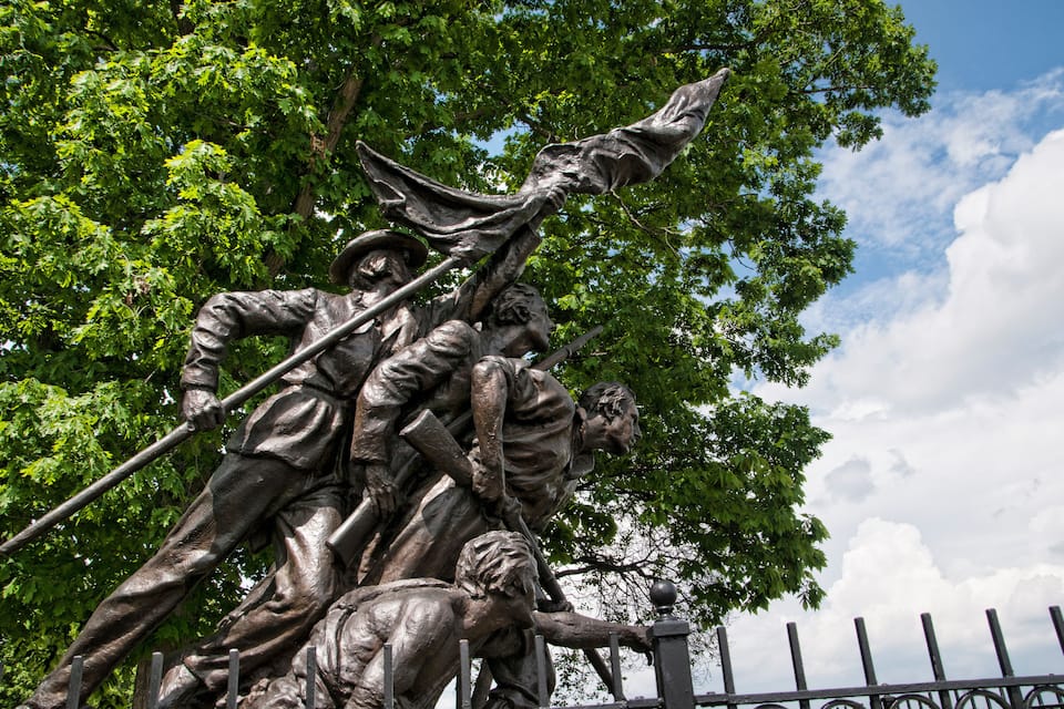 Una estàtua de bronze de soldats que pugen una collada, envoltada d'arbres i un cel serè.