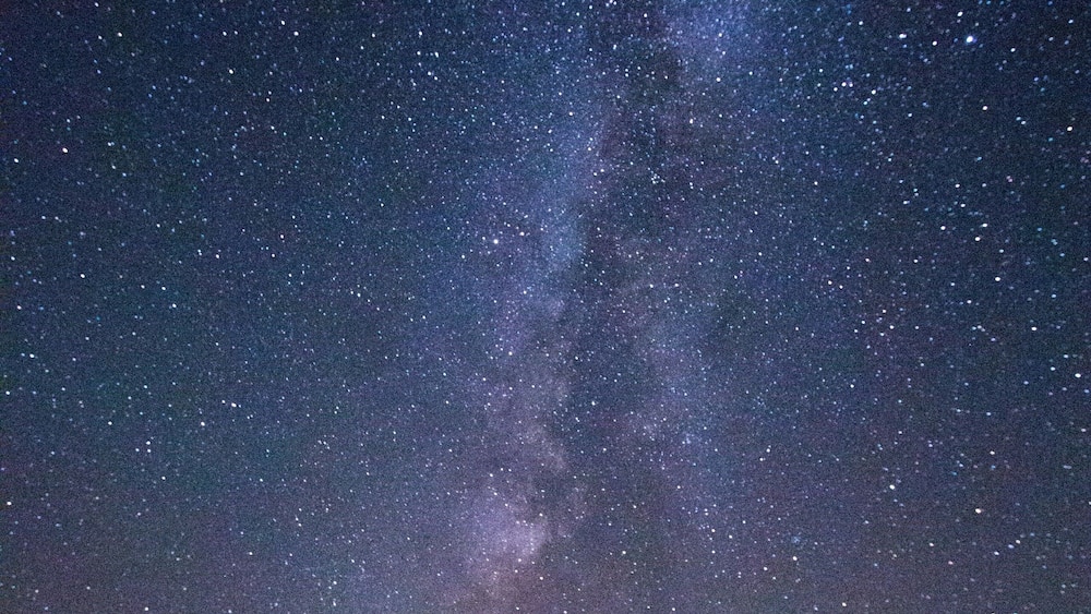 A starry night sky with the Milky Way visible, a forested hill, and two tents.