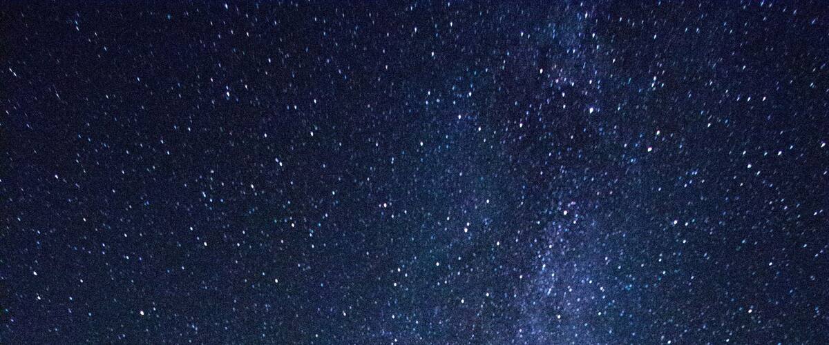 A starry night sky with the Milky Way visible, a forested hill, and two tents.
