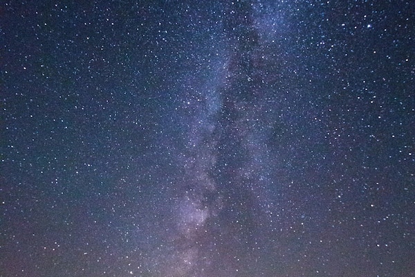Un ciel étoilé où l’on peut apercevoir la Voie lactée, une colline boisée et deux tentes.