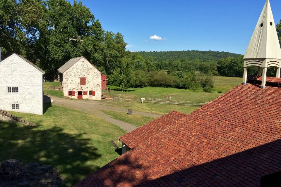 Un paysage rural avec une grange en pierre, un bâtiment à toit rouge et le clocher d’une église.
