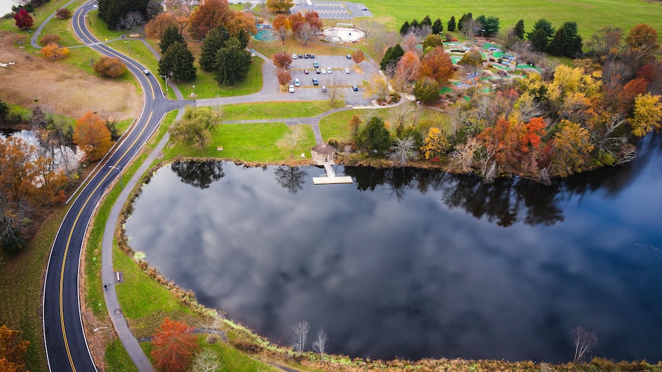 An aerial view of a park with a winding road, a lake, and autumn-colored trees.