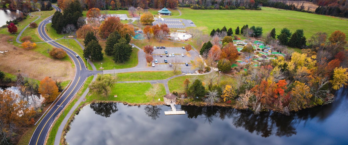 An aerial view of a park with a winding road, a lake, and autumn-colored trees.