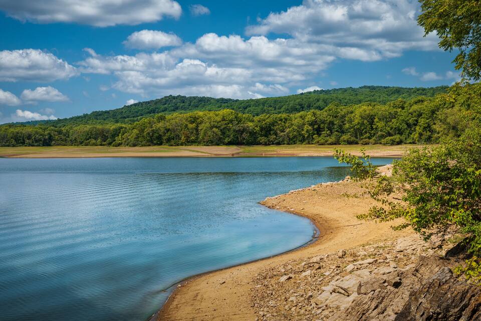 A calm lake with a sandy shoreline and a forested hill in the background.