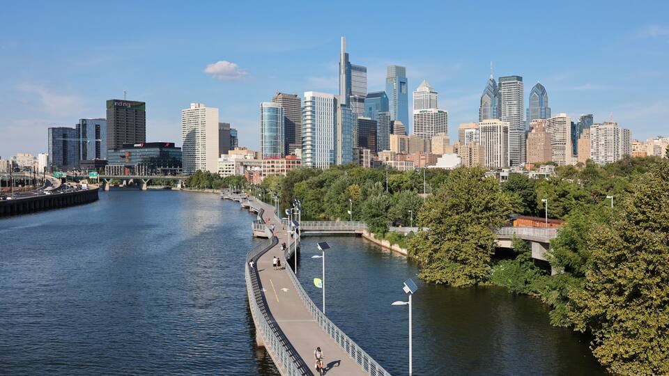 A river with a pedestrian bridge, a city skyline, and a park with trees.