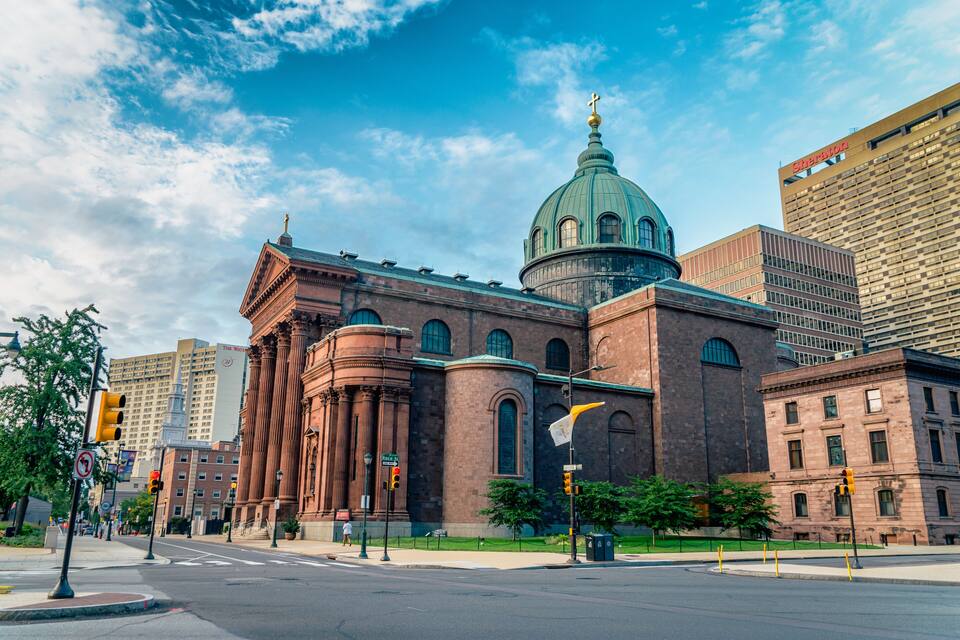 Une église historique dotée d’un dôme et de colonnes, entourée de bâtiments modernes et surplombée d’un ciel dégagé.