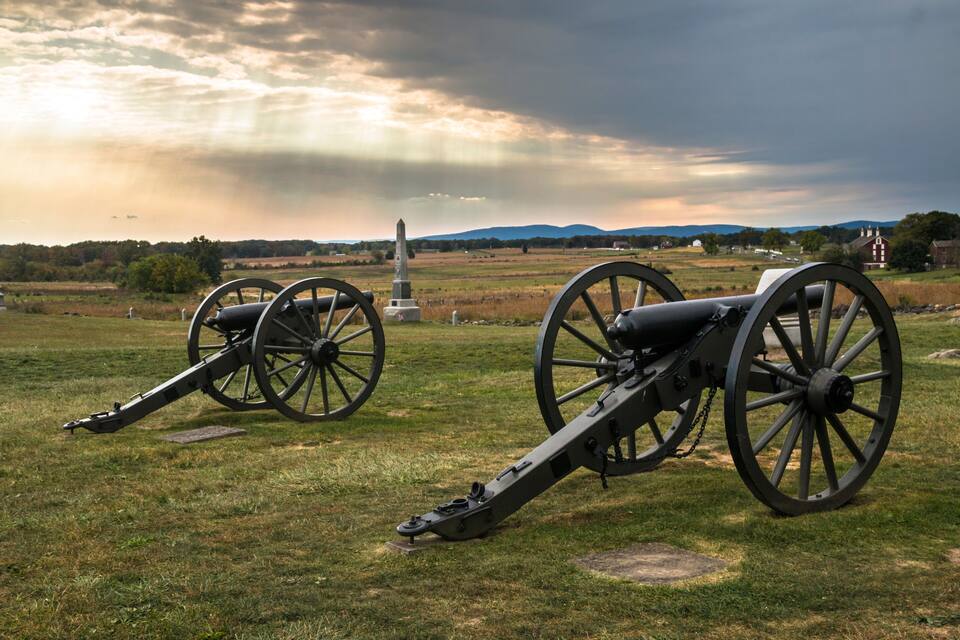 Two cannons in a grassy field with a cloudy sky and distant mountains.