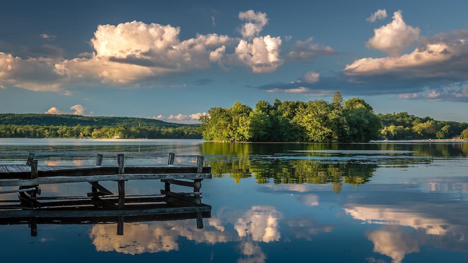 A calm lake with a wooden dock, surrounded by dense green forest and a hill in the background.