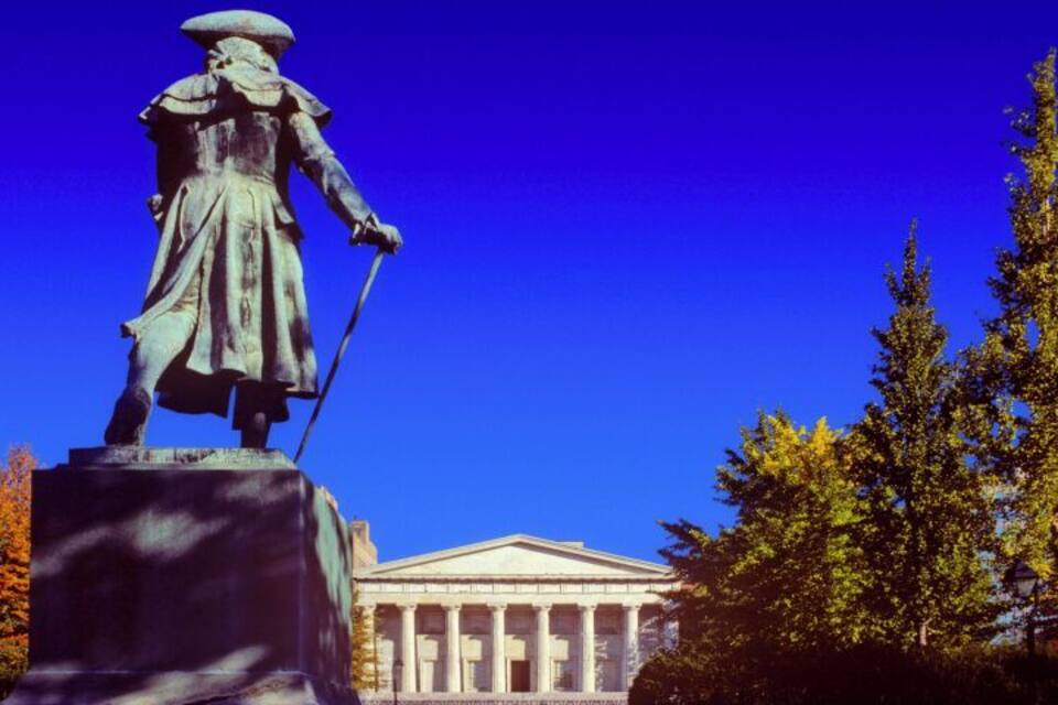A statue of a historical figure, a neoclassical building with columns, and autumn foliage.