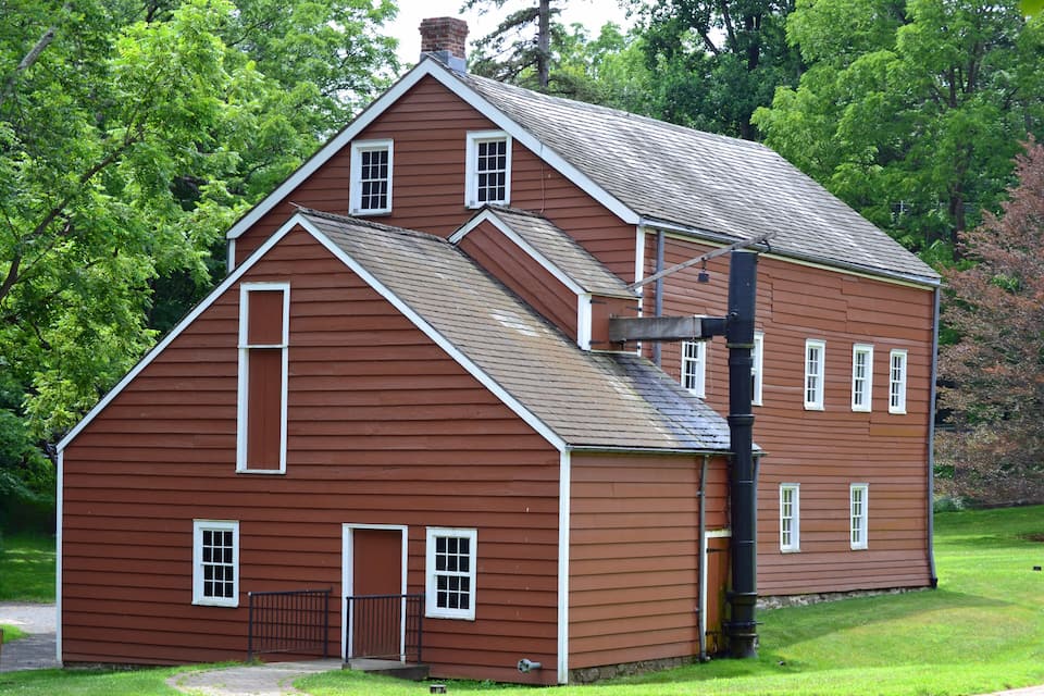 A large, two-story wooden house with a steep gable roof, surrounded by lush green trees.