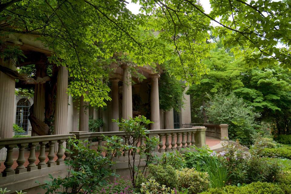 A classical building with columns and a balcony, surrounded by lush greenery and a well-maintained garden.