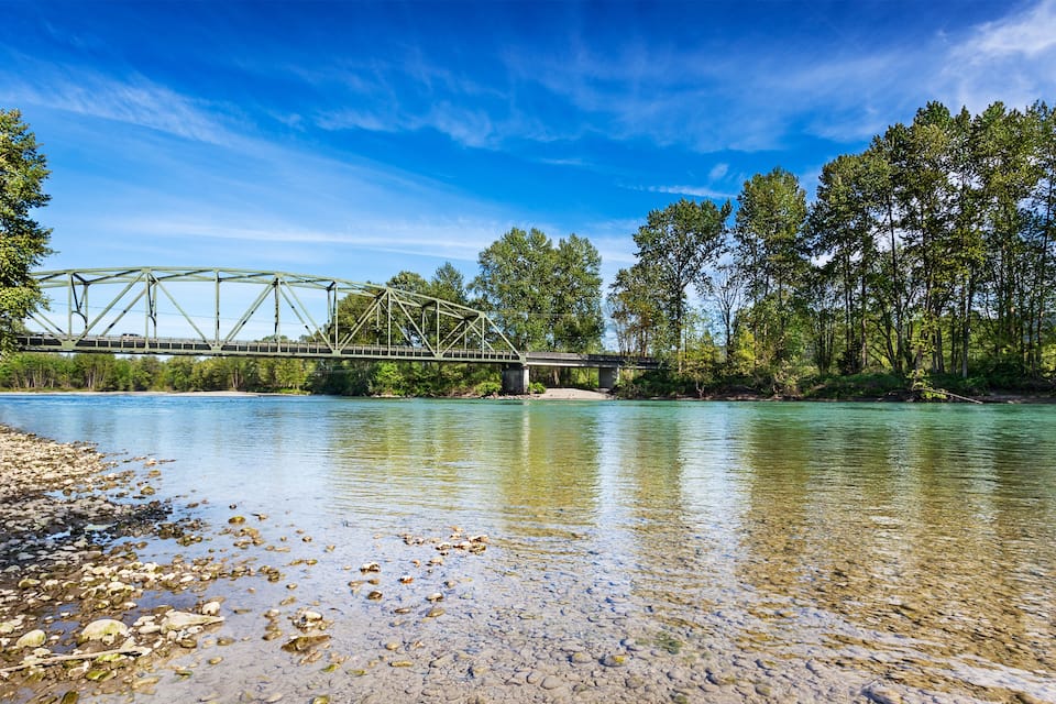 A green metal bridge over a river with clear water, surrounded by trees and pebbly shore.