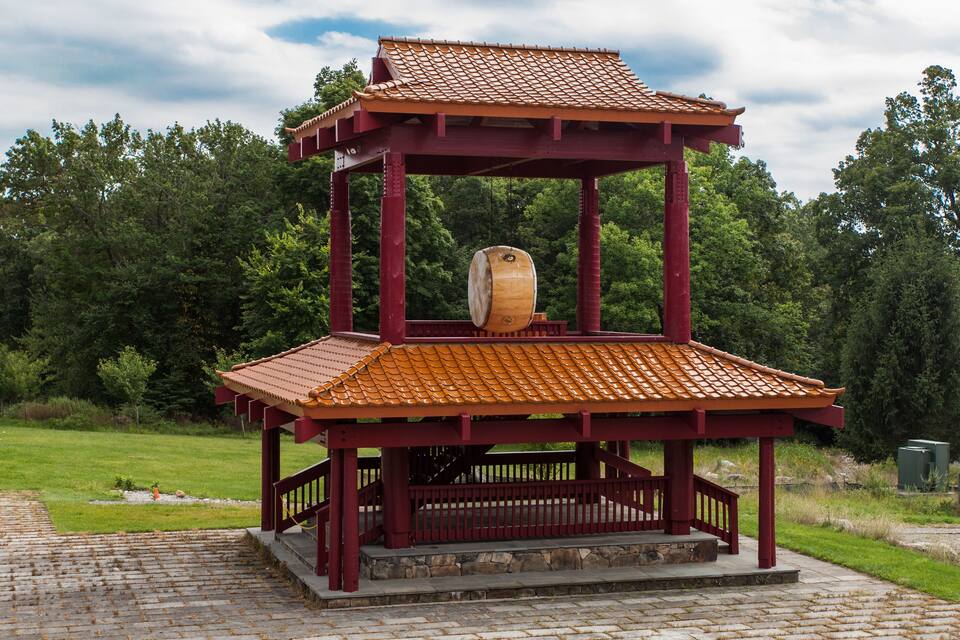A traditional wooden gazebo with a tiled roof and a barrel in the center, surrounded by a grassy area and trees.