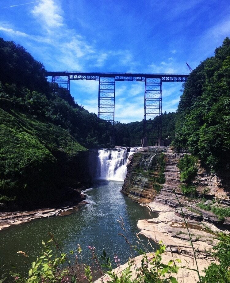 A waterfall under a bridge, surrounded by lush greenery.