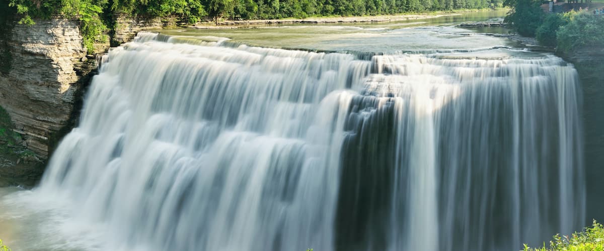 A large waterfall cascading down a rocky cliff into a river surrounded by greenery.