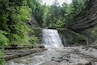 Ein Wasserfall stürzt in einen Fluss, umgeben von dichtem Wald und steilen Felsen.