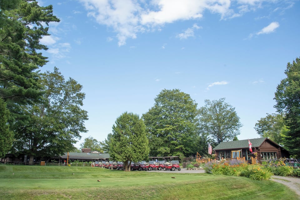 A golf course with a clubhouse, a row of parked cars, and a large tree in the foreground.