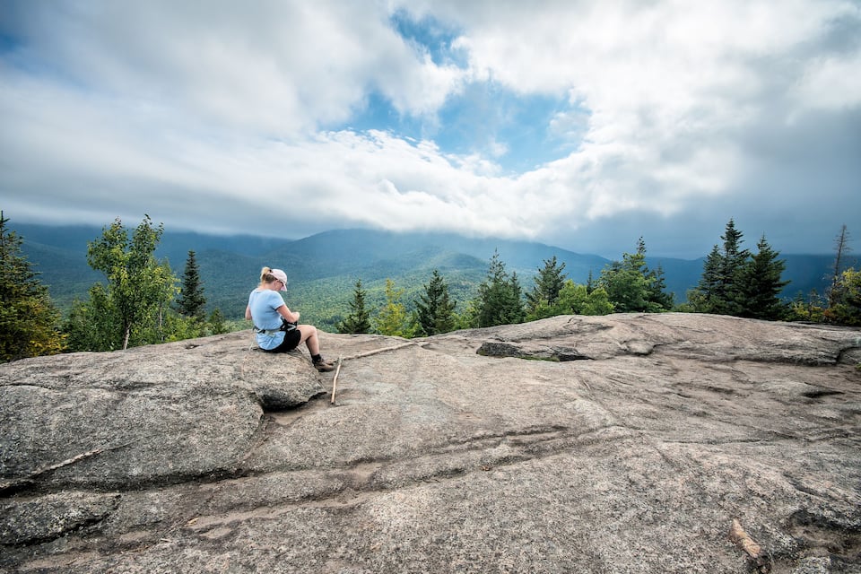A person sitting on a large rock with a mountainous landscape in the background.