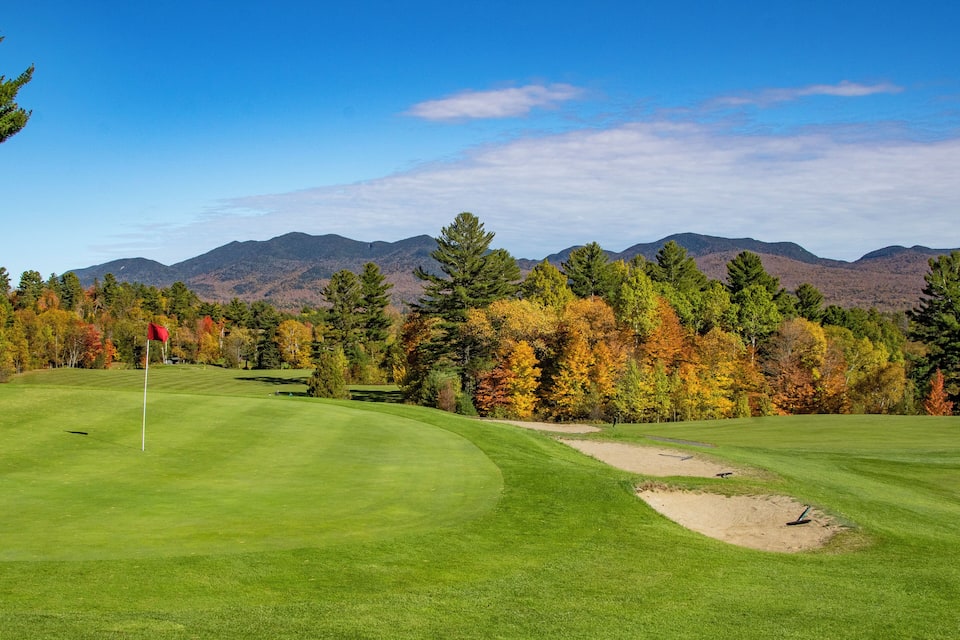 A golf course with a red flag, sand bunkers, and autumn-colored trees in the background.