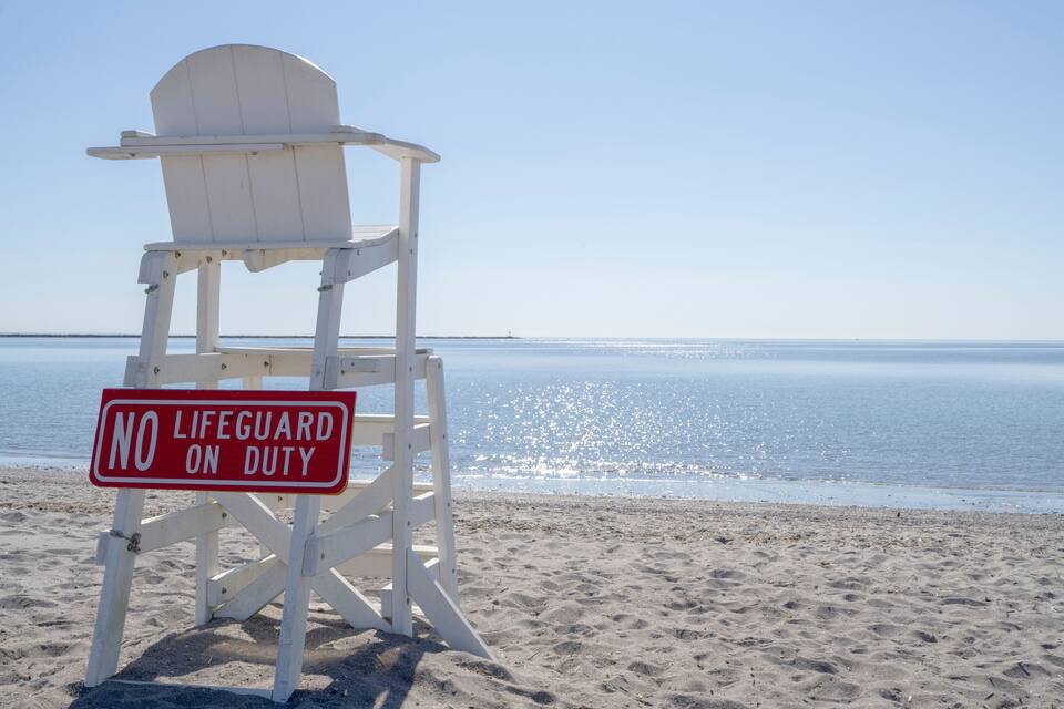 A lifeguard chair with a sign that reads "NO LIFEGUARD ON DUTY" on a beach with calm water and a clear sky.