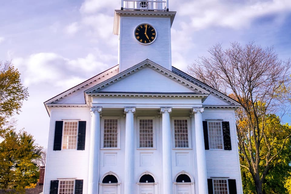 A white church with a steeple and a clock, surrounded by trees and a partly cloudy sky.