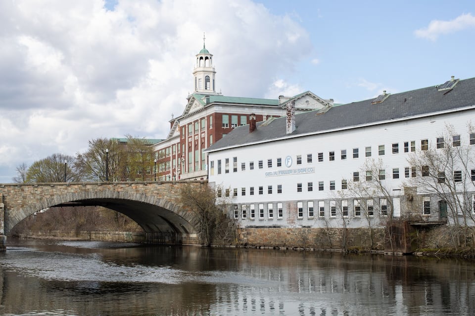 A stone bridge over a river, a white building with "GEO. FULLER & SON CO." on it, and a red building with a tower in the background.