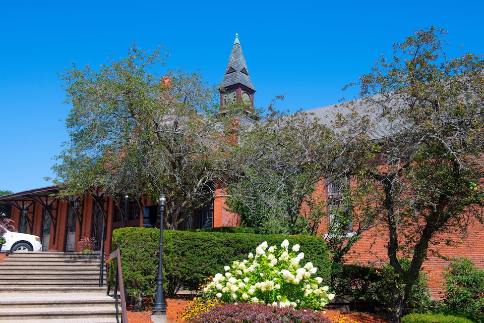A brick building with a clock tower, surrounded by trees and a flower bed.