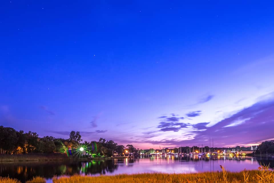 A nighttime lakeside scene with a starry sky, a calm lake reflecting lights, and a distant city skyline.