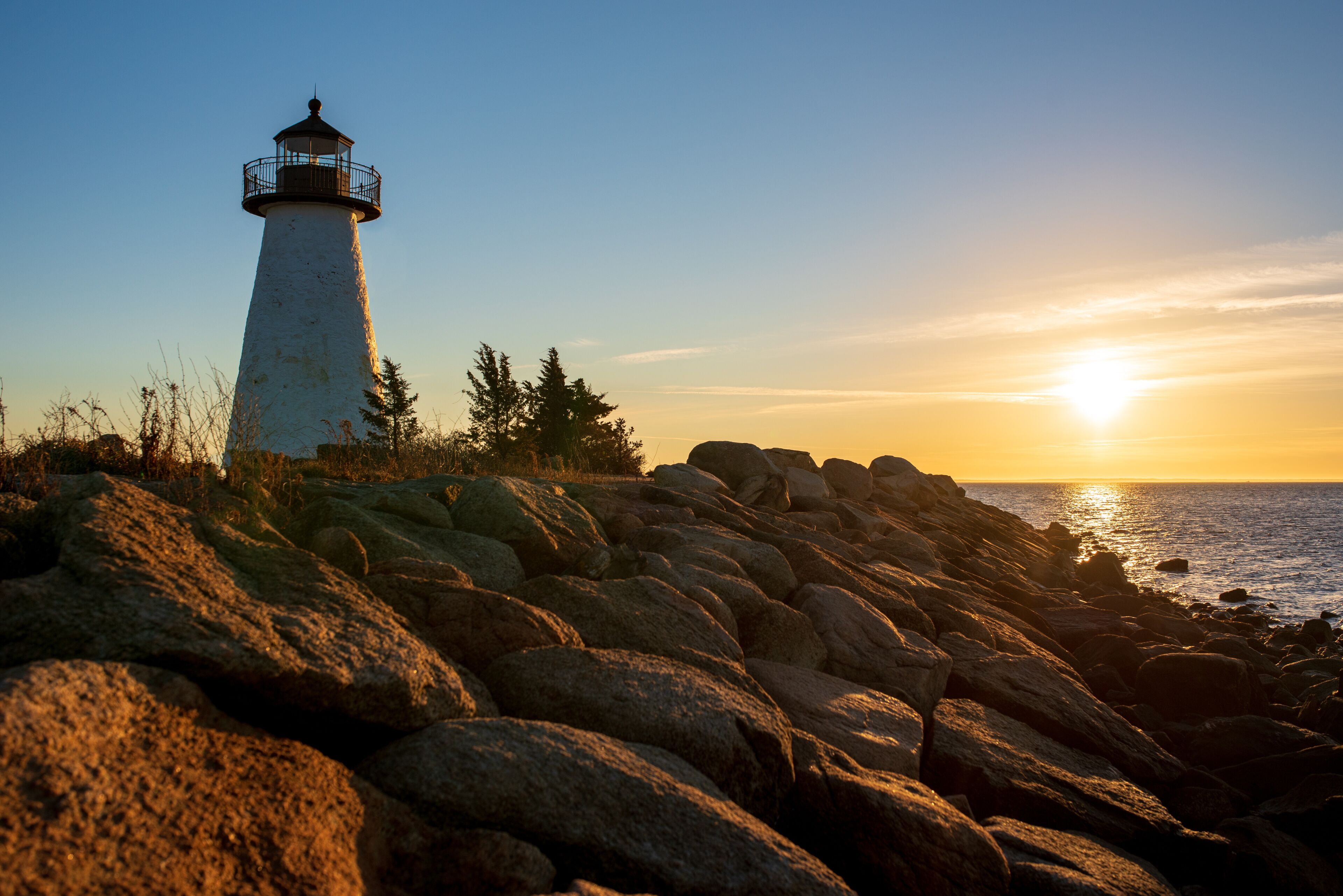 A lighthouse on a rocky shore with the sun setting in the background.