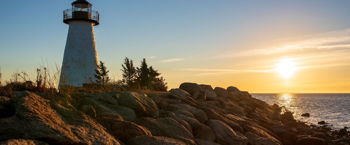 A lighthouse on a rocky shore with the sun setting in the background.