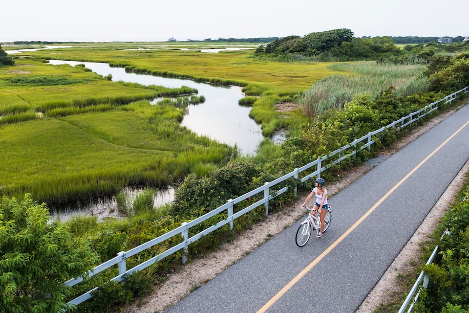 A cyclist on a paved path alongside a river with marshland and a fence.