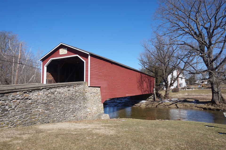 A red covered bridge over a small stream, surrounded by trees and a stone wall.