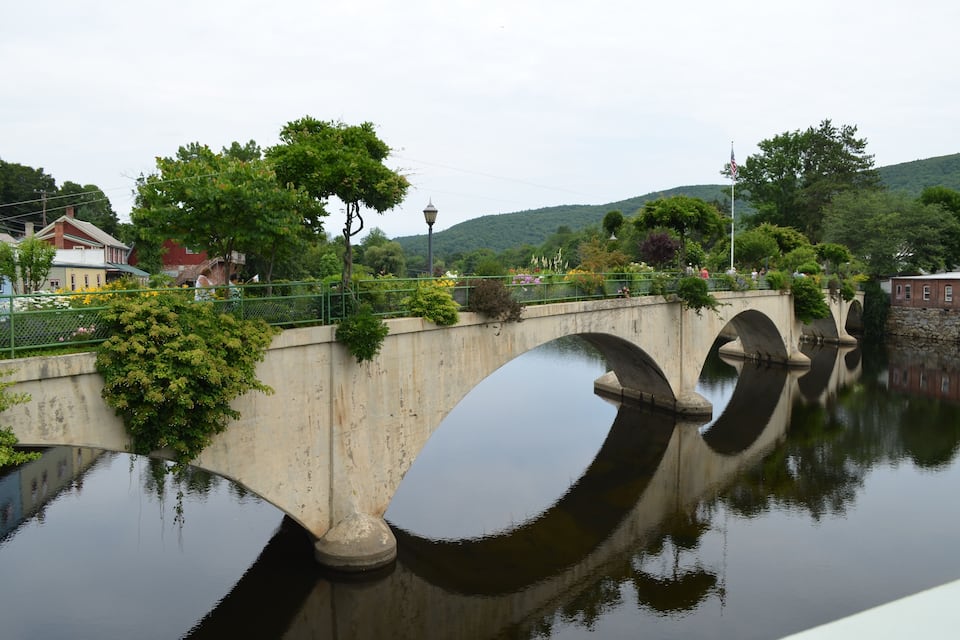 A concrete bridge with three arches over a calm river, surrounded by greenery and buildings.