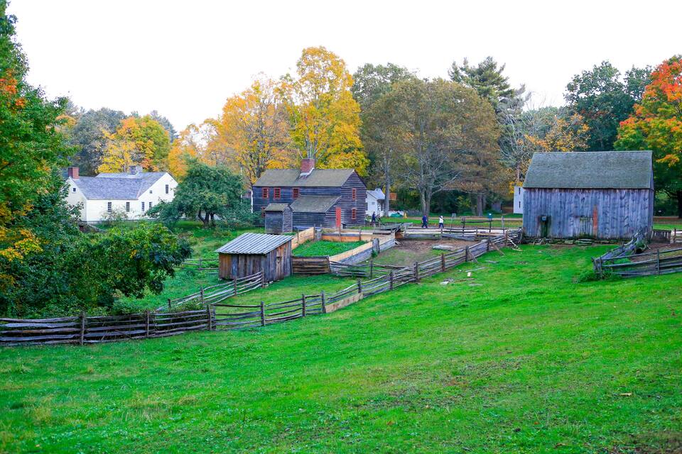 A rural landscape with a white house, a wooden barn, and a fenced area with trees displaying autumn foliage.