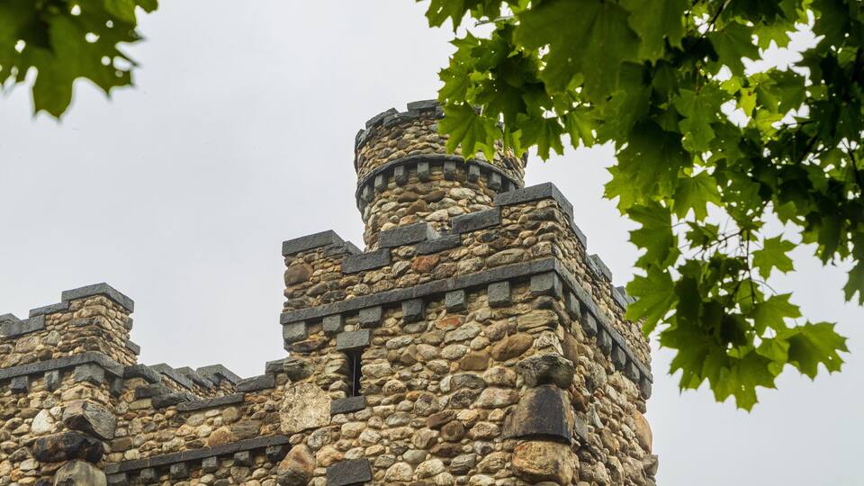 Ein steinerner Turm mit einer runden Spitze und einem rechteckigen Fundament, umgeben von Bäumen.