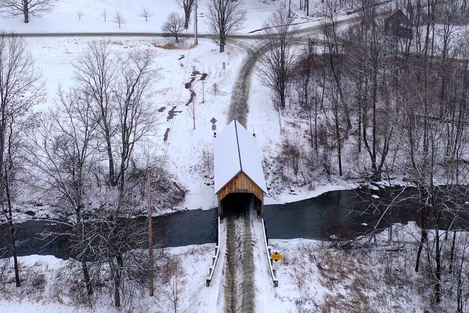 A snow-covered landscape with a wooden covered bridge over a river, surrounded by trees and a snowy path.