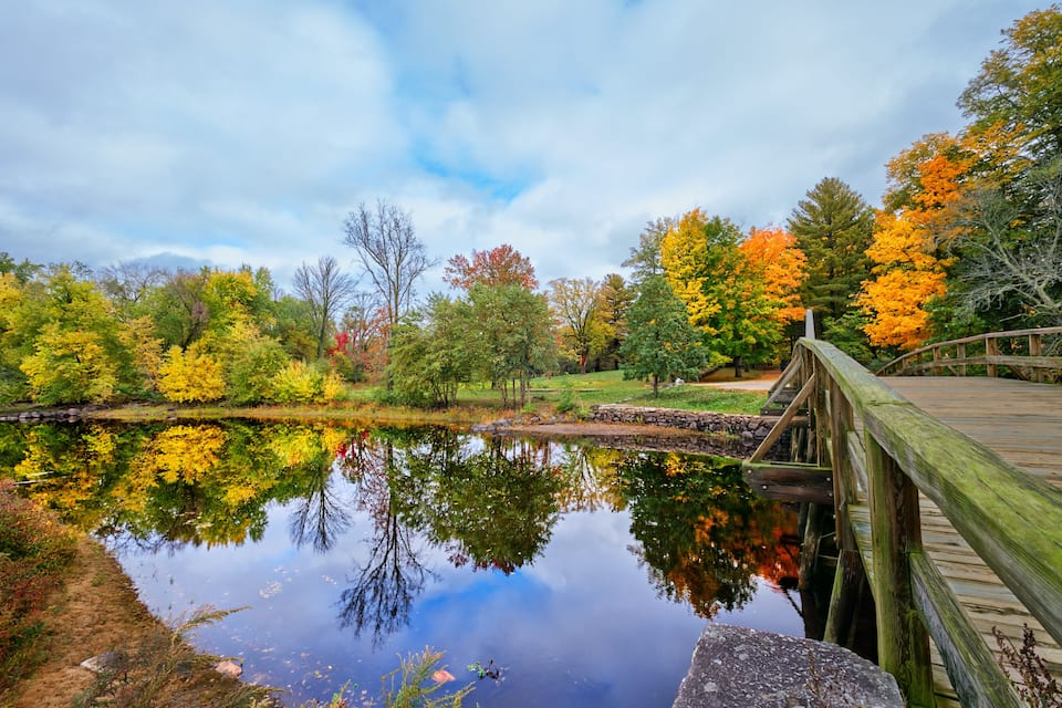 Un pont en bois enjambant un lac paisible, entouré d’arbres aux couleurs automnales.