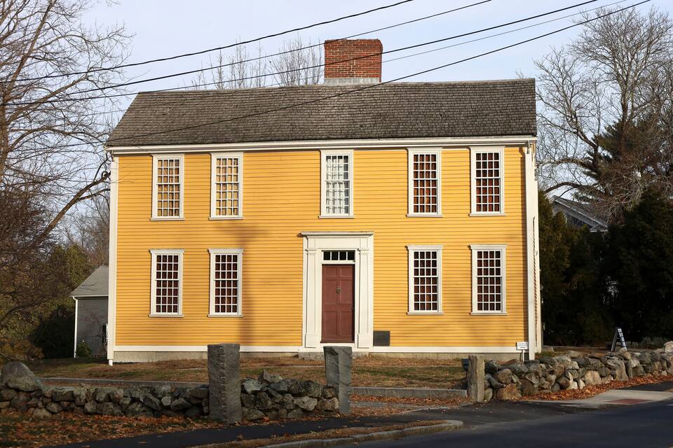 A yellow house with a brown door and white trim, surrounded by a stone wall and a stone pillar.