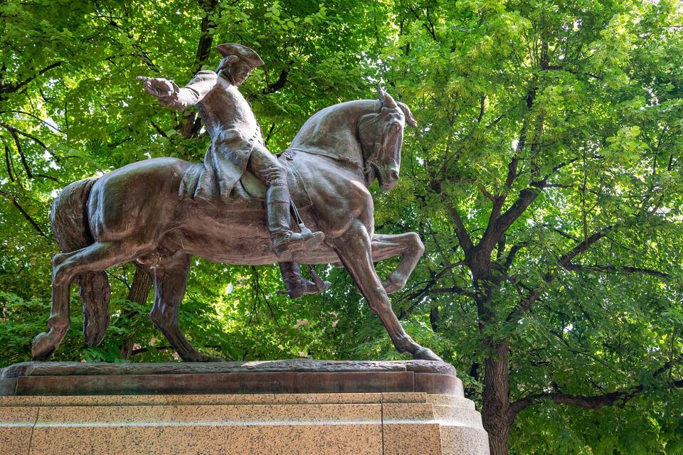 Estátua de bronze de um homem a cavalo, sobre um pedestal de pedra, rodeada por árvores verdes e exuberantes.