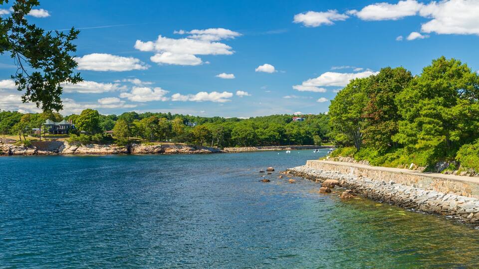 A calm lake with a stone wall and trees on the right, a house on the left, and a clear blue sky.