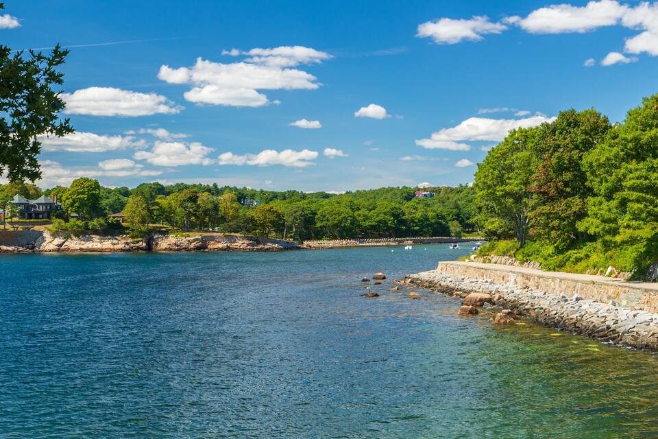 A calm lake with a stone wall and trees on the right, a house on the left, and a clear blue sky.