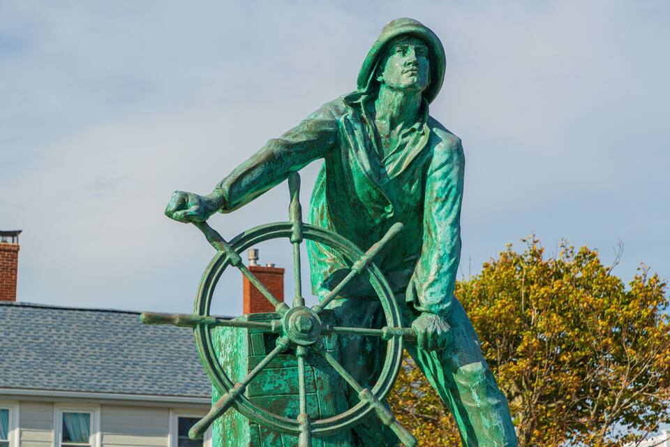 A statue of a person with a wheel, set against a backdrop of houses and a tree.