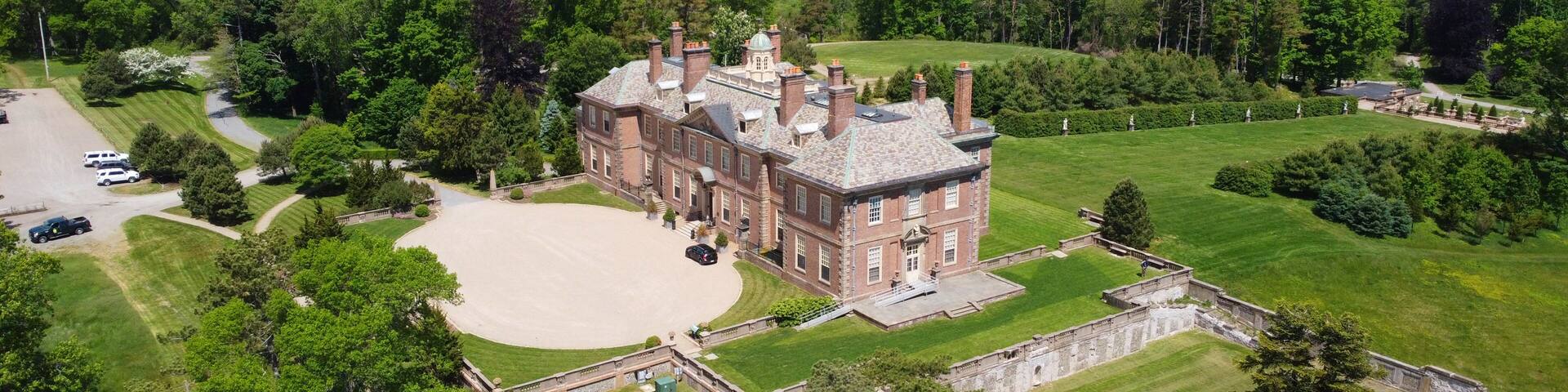 A large estate with a prominent brick building, expansive lawns, and a body of water in the background.