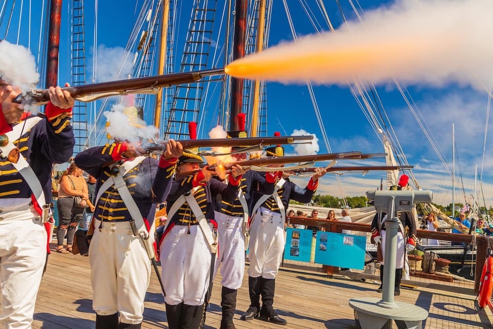 Un grupo de personas con uniformes navales en una cubierta de madera, con aparejo de barco y un cielo azul despejado al fondo.
