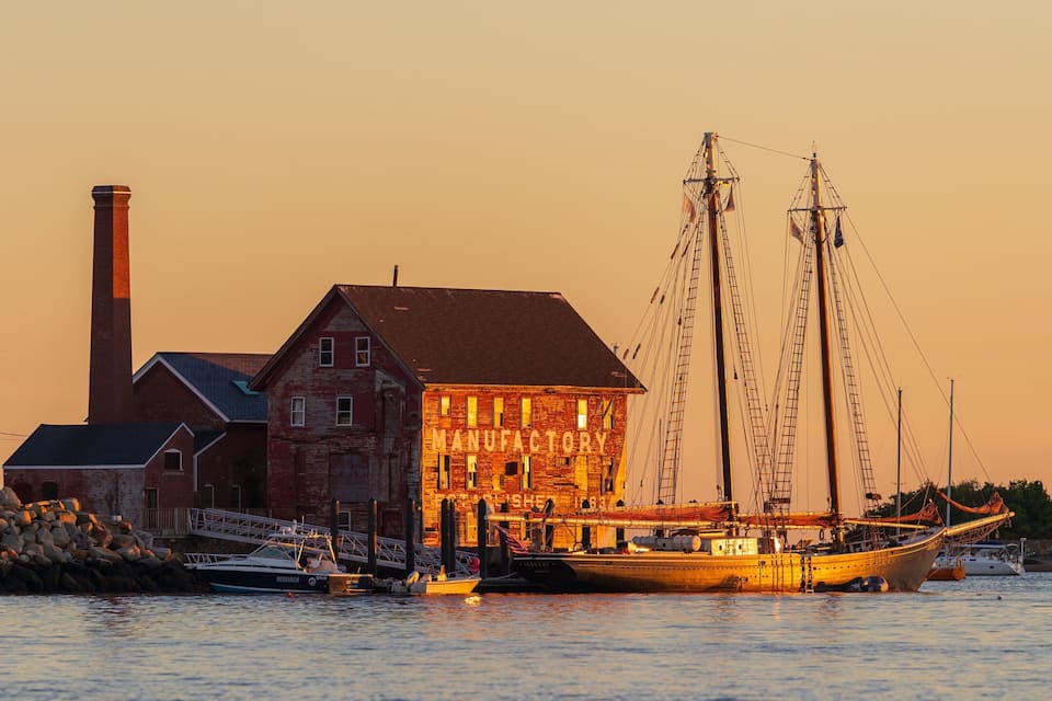 A historic building labeled "MANUFACTURY" with a large chimney, a dock with fishing boats, and a sunset sky.