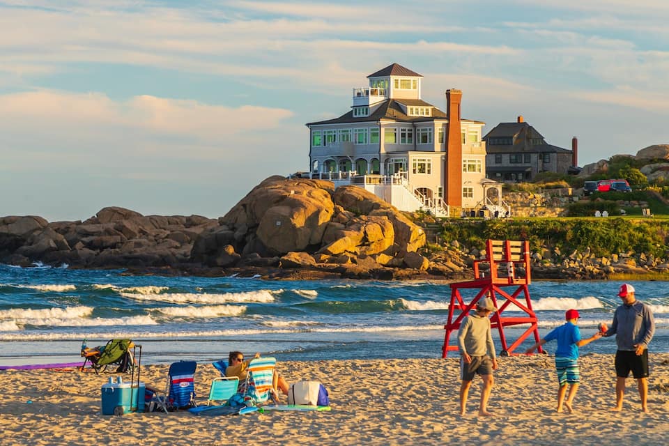 Una playa con una torre de salvavidas, olas y una casa grande con torre sobre una costa rocosa.