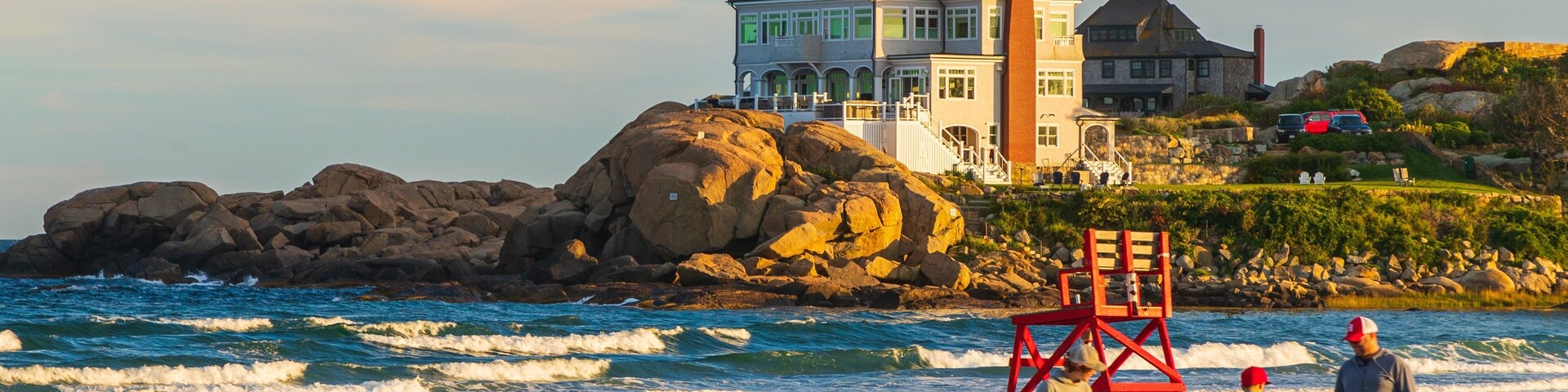 A beach with a lifeguard tower, waves, and a large house with a tower on a rocky shore.