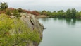 A rocky shoreline with calm water, surrounded by trees with autumn foliage.