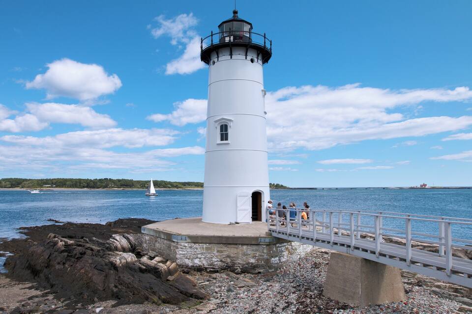Um farol branco com topo preto, localizado numa costa rochosa com céu azul límpido e um veleiro ao fundo.