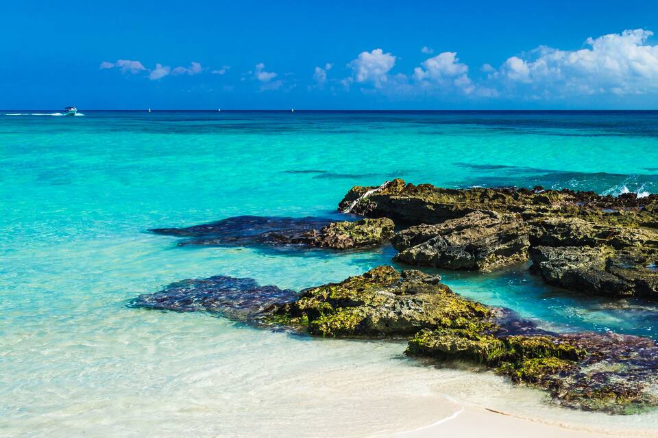 Une mer turquoise aux eaux limpides, une plage de sable blanc et des affleurements rocheux couverts d’algues.